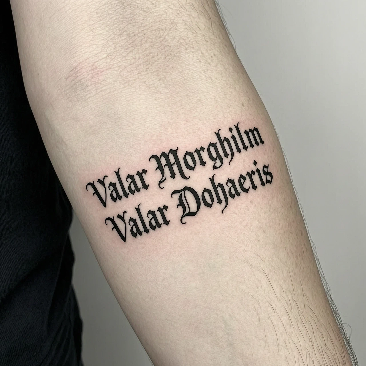 Татуировка на коже предплечья: две строки текста на высоком валирийском «Valar Morghulis» и «Valar Dohaeris». Шрифт строгий, каллиграфический, будто выгравирован на металле.…