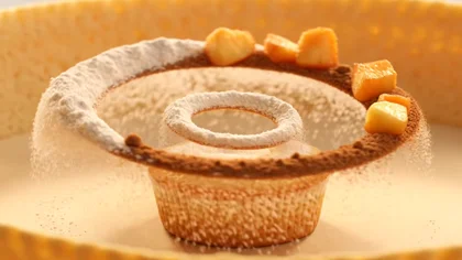 Flour, sugar, cinnamon, and apple pieces float in midair, spinning in a slow-motion spiral. The ingredients merge and form a cupcake covered in glossy glaze and powdered sugar.…